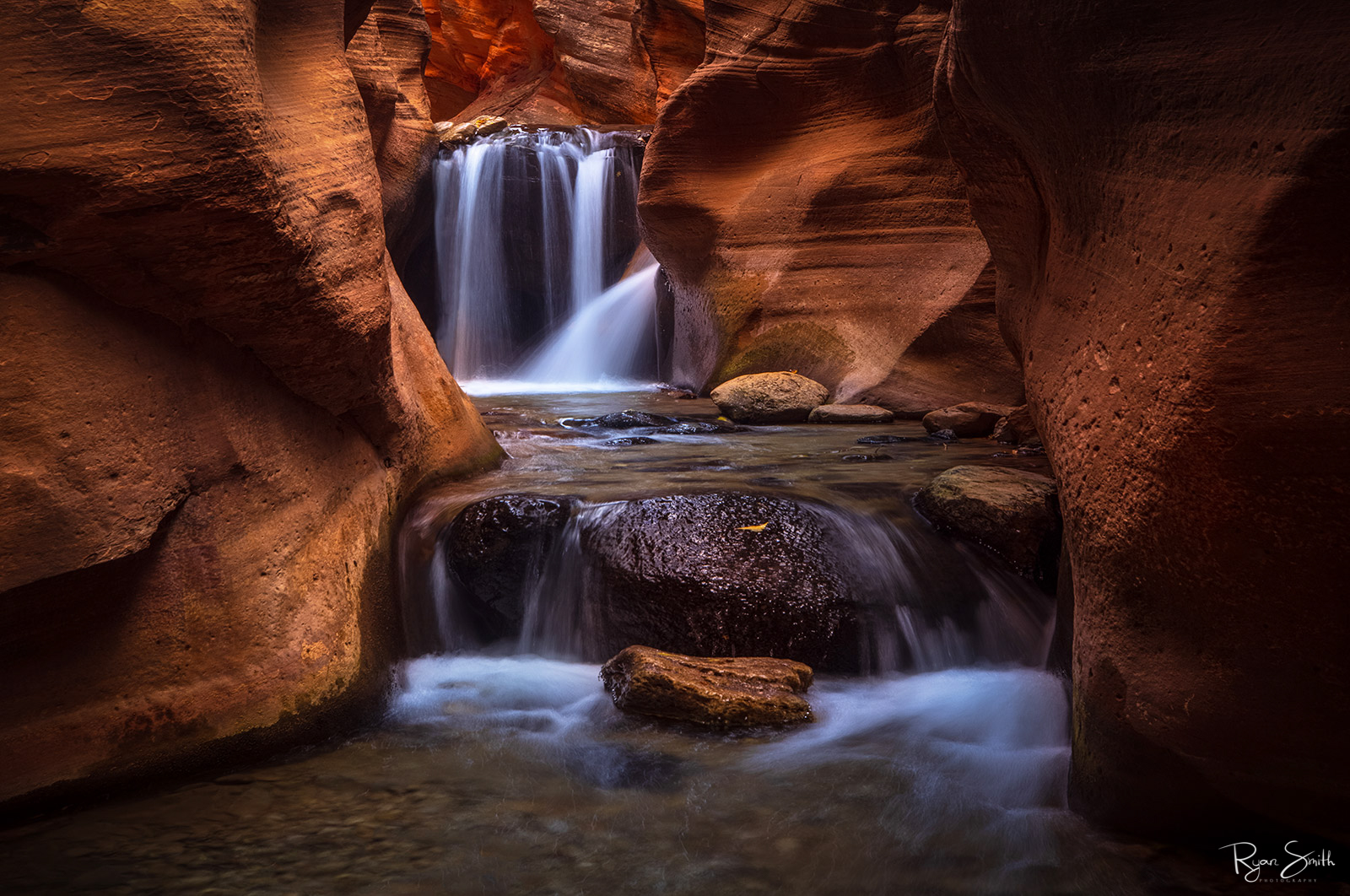 A waterfall in a sandstone slot canyon with reddish-orange walls and two levels of the waterfall.