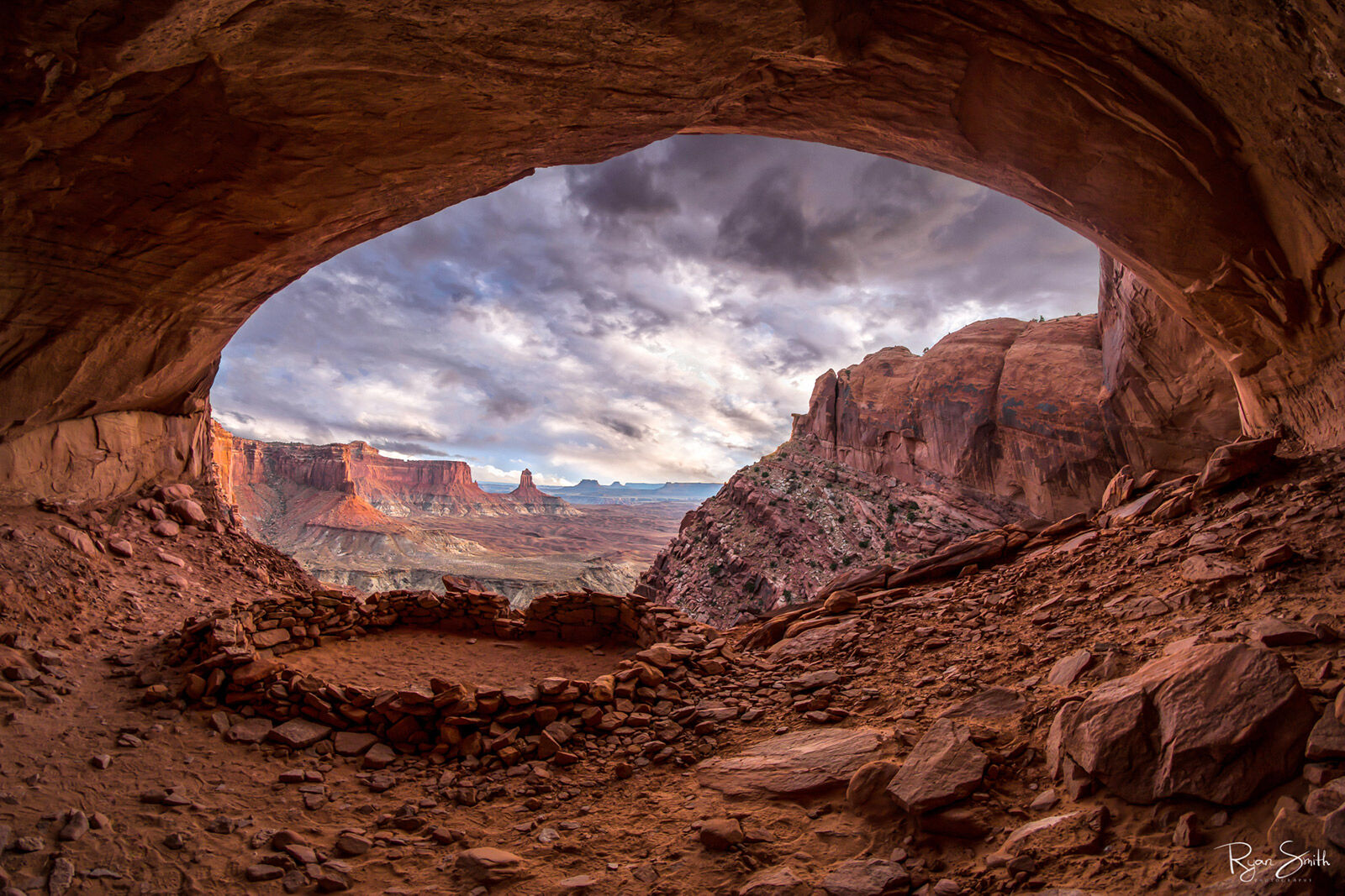 A circle of stones used for ancient religious purposes sits in a cave like kiva with a sky full of white clouds and a blue sky.