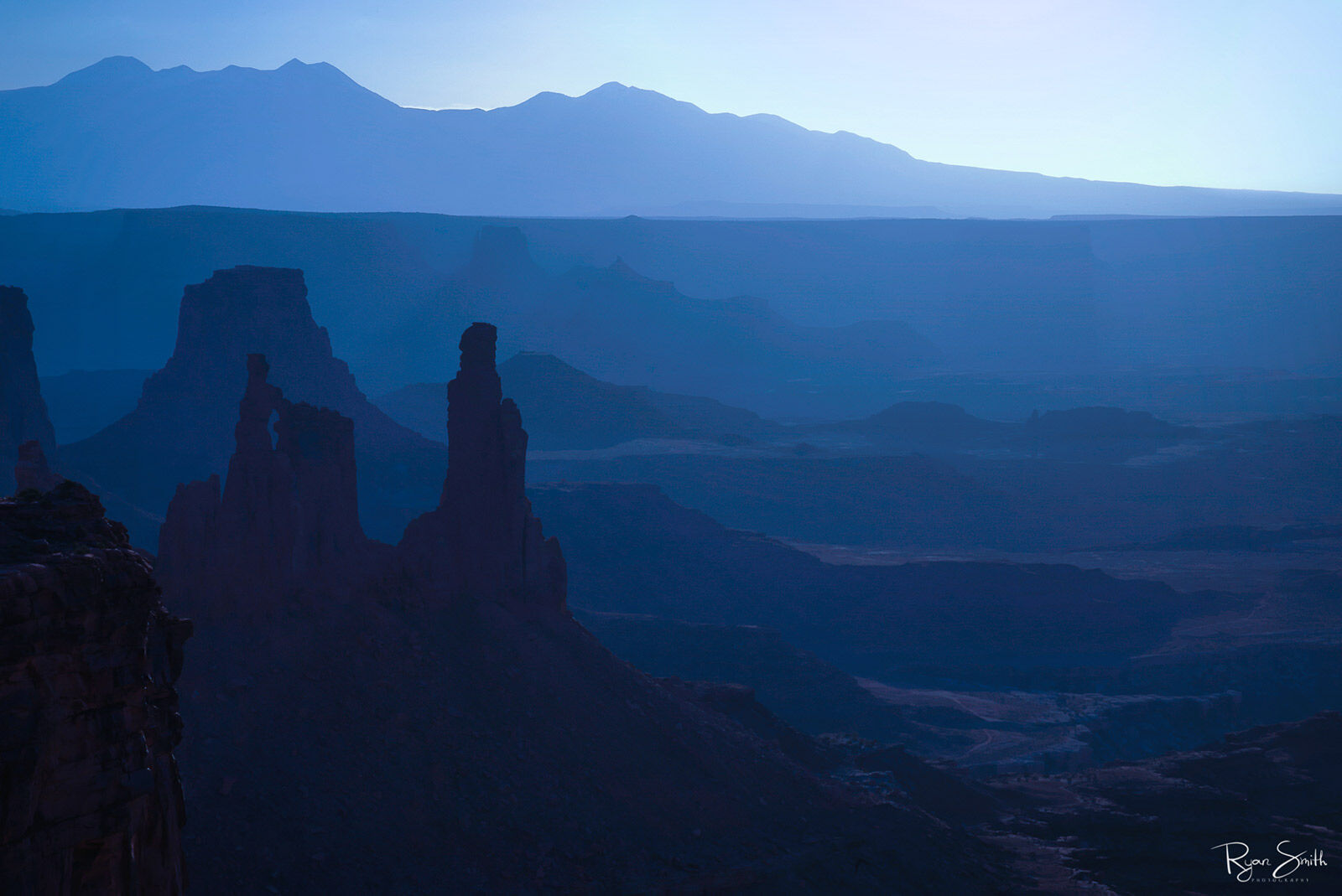 With the sun below the horizon at sunrise, a desert landscape shows hues of blue with first light and blue silhouettes of the rock formations and sky line.