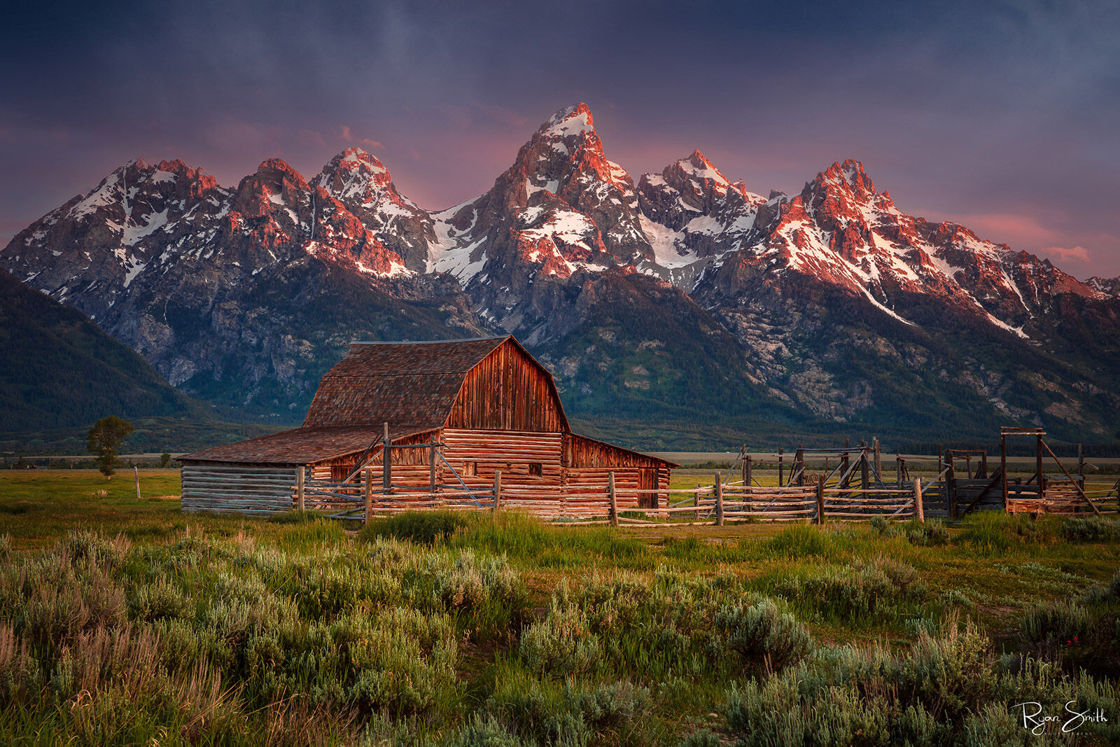A rustic barn with a wooden fence around it sits at the foot of a grand mountain range at sunrise. Pink clouds peek from behind mountains kissed with sunlight.