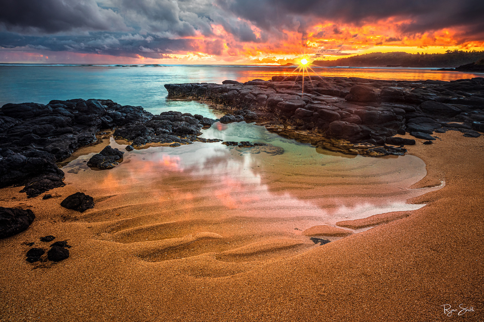 The sand in a small tide pool holds a wave-like pattern while along the rocky shore aqua blue water gently comes in and on the right reflects the orange sunset.