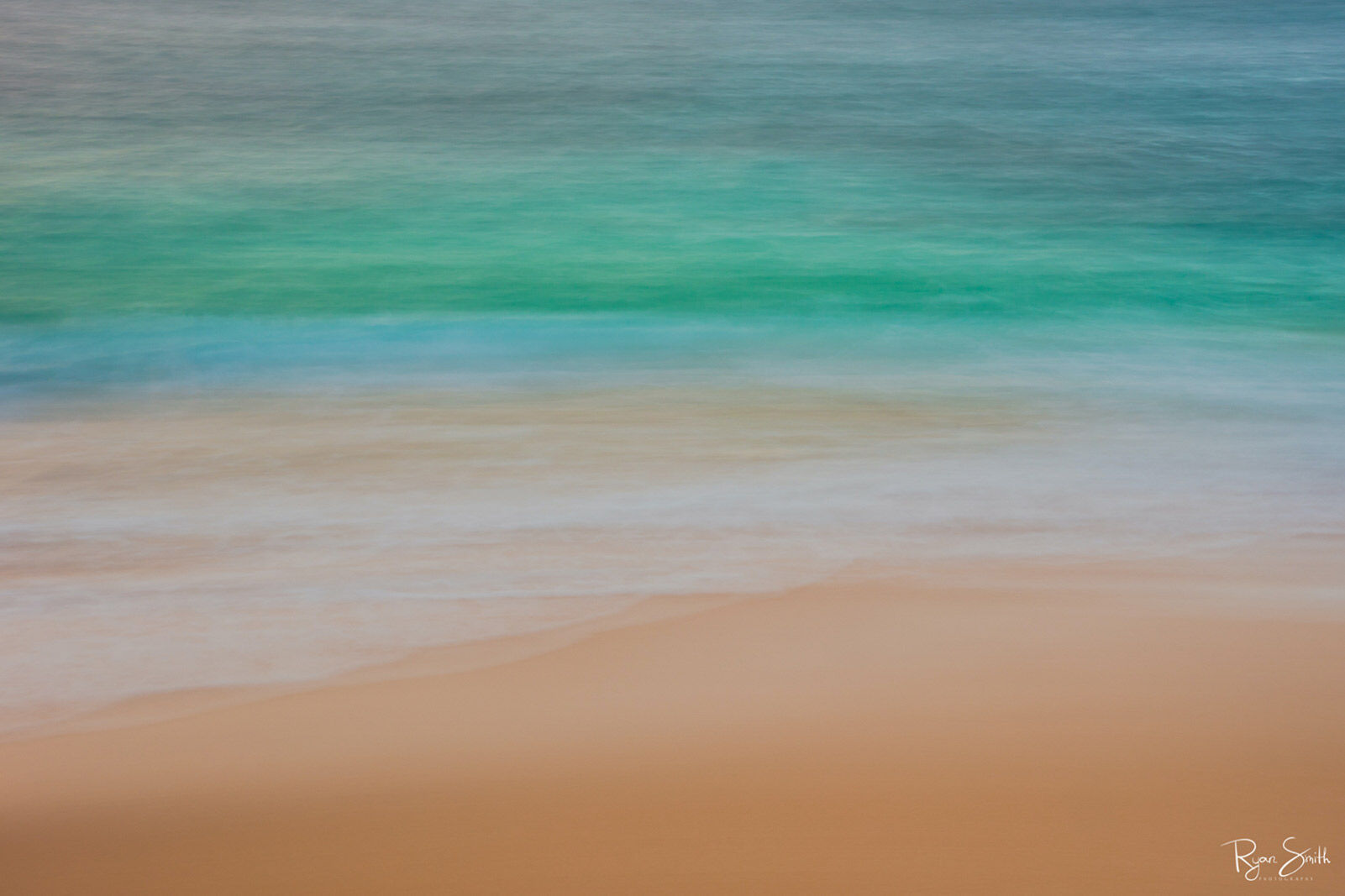 Abstract photography of the beach close up with the tan sand, shallow water and deep turquoise and blue water out further.